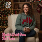 Woman in green Joy hoodie with red yarn, sitting by Christmas tree and fireplace, cozy holiday setting.