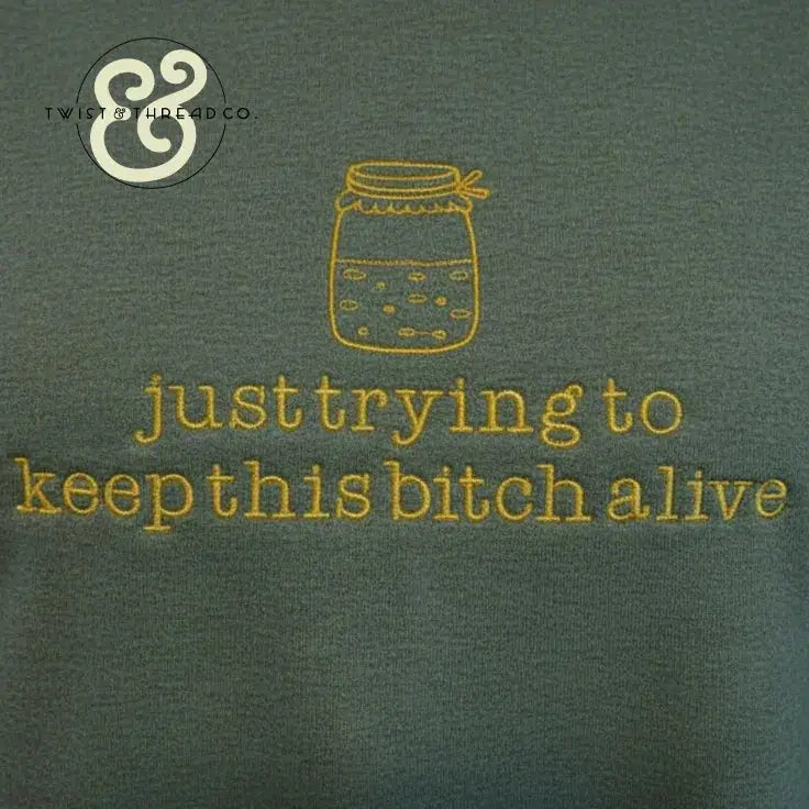 Green fabric with yellow embroidered jar and text: just trying to keep this bitch alive, casual graphic apparel