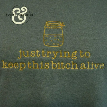 Green fabric with yellow embroidered jar and text: just trying to keep this bitch alive, casual graphic apparel