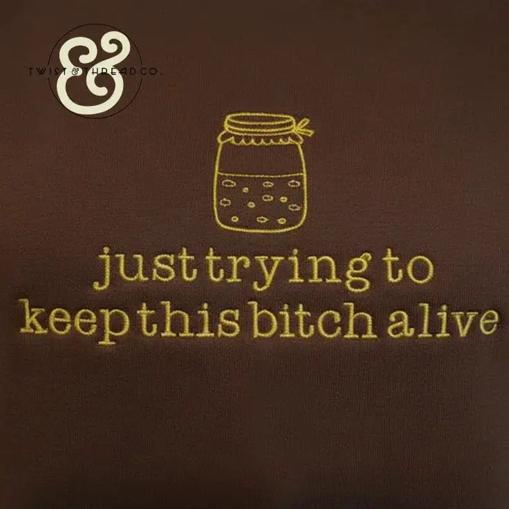 brown fabric with yellow embroidery of a jar and text saying just trying to keep this bitch alive
