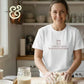 Smiling woman kneading dough in kitchen wearing white t-shirt with mason jar and text 'just trying to keep this bitch alive'
