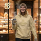 Smiling woman in beige hoodie and knit hat holding coffee cup outside bakery at night