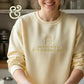 Smiling woman wearing cream sweatshirt with embroidered jar and text 'just trying to keep this bitch alive' in kitchen