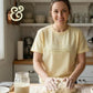 Smiling woman kneading dough in bright kitchen wearing light yellow shirt with humorous text