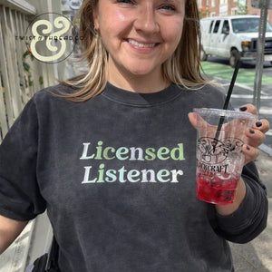 Smiling woman wearing a black sweatshirt with embroidered text 'Licensed Listener' holding a clear drink cup outdoors