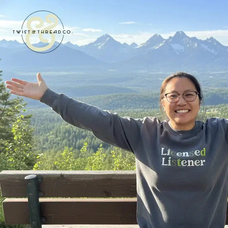 Smiling woman wearing a Licensed Listener embroidered sweatshirt outdoors with mountain landscape backdrop
