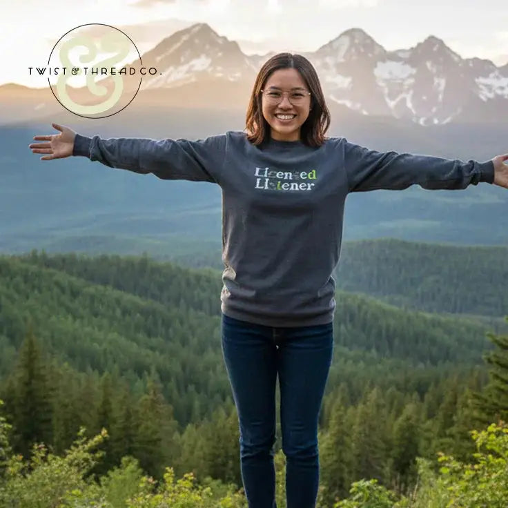 Smiling woman outdoors wearing a dark embroidered sweatshirt reading 'Licensed Listener' with mountains and forest background