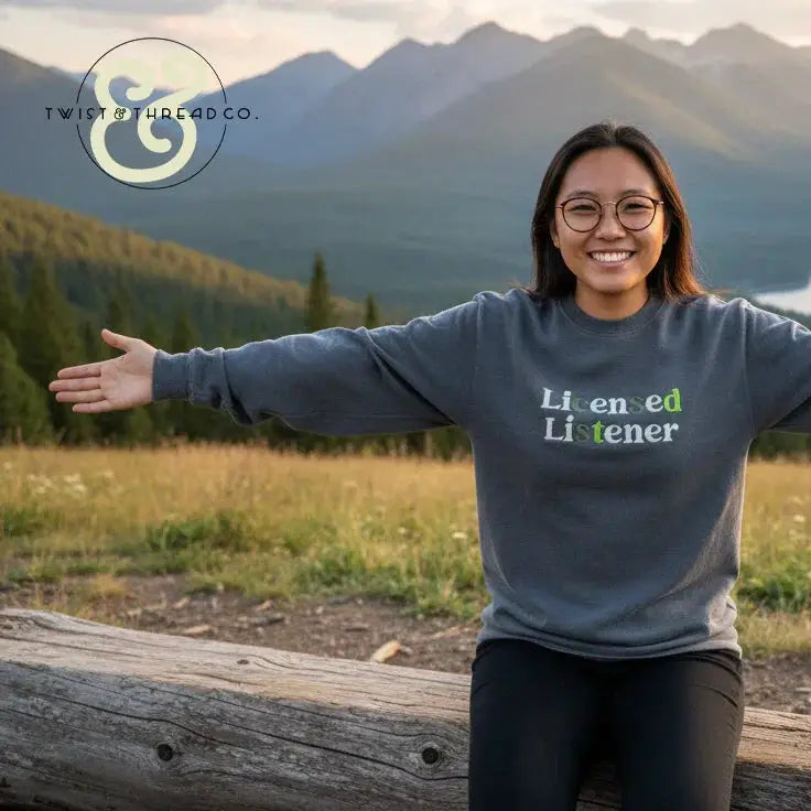 Smiling woman wearing a grey 'Licensed Listener' embroidered sweatshirt by Twist & Thread Co., outdoors with mountain landscape
