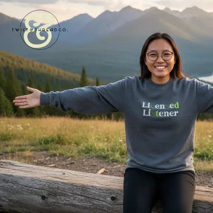 Smiling woman wearing a grey 'Licensed Listener' embroidered sweatshirt by Twist & Thread Co., outdoors with mountain landscape