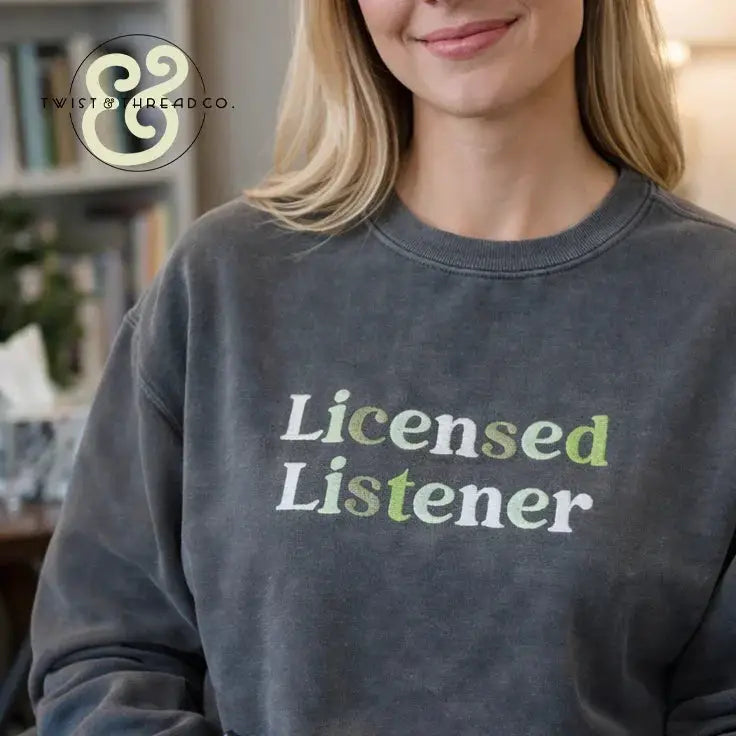 Smiling woman wearing a charcoal grey embroidered sweatshirt with 'Licensed Listener' text in white and green, indoor setting