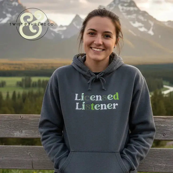Smiling woman wearing a gray hoodie with 'Licensed Listener' embroidery, outdoors with mountain backdrop