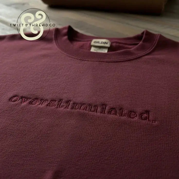 Maroon Gildan sweatshirt with embroidered word 'overstimulated' on chest, laid on wooden surface