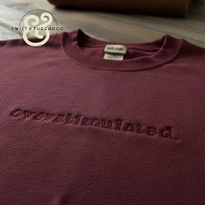 Maroon Gildan sweatshirt with embroidered word 'overstimulated' on chest, laid on wooden surface