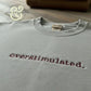 Light grey Gildan sweatshirt with burgundy embroidered text 'overstimulated.' on wooden surface