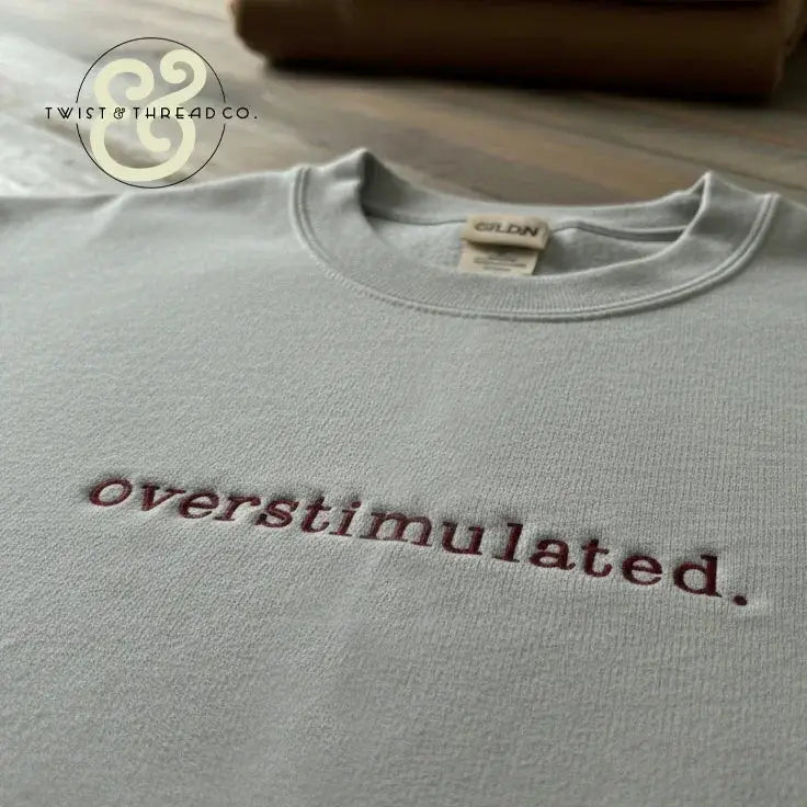 Light grey Gildan sweatshirt with burgundy embroidered text 'overstimulated.' on wooden surface
