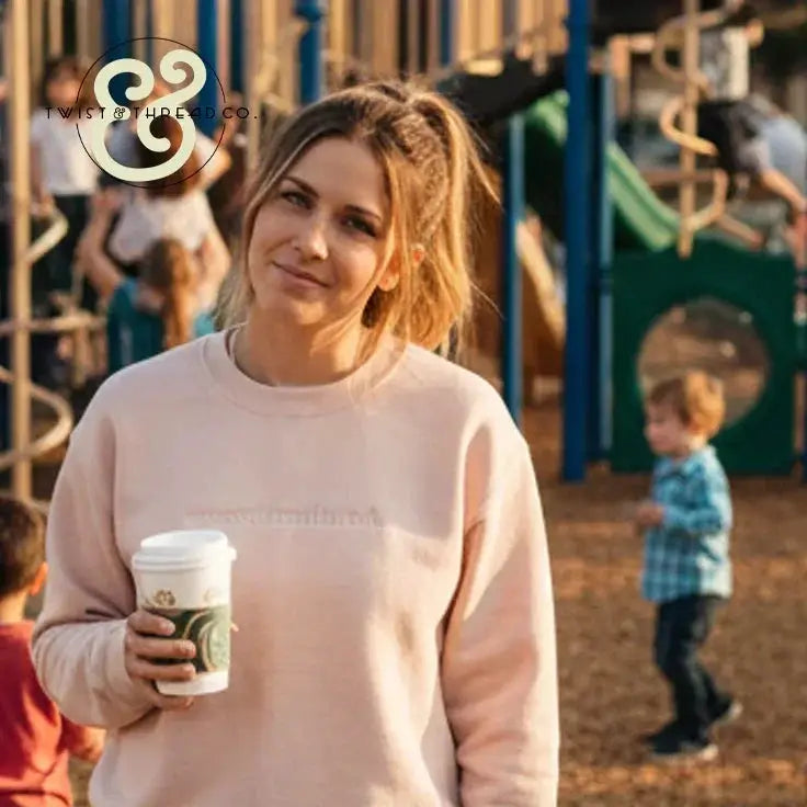 Woman wearing blush pink sweatshirt holding coffee cup at sunny playground with children