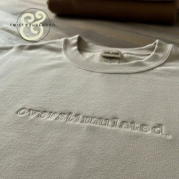 Close-up of a white Gildan sweatshirt with embroidered word 'overstimulated' on a wooden surface