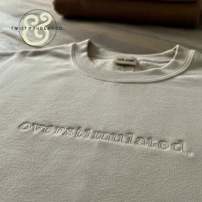 Close-up of a white Gildan sweatshirt with embroidered word 'overstimulated' on a wooden surface