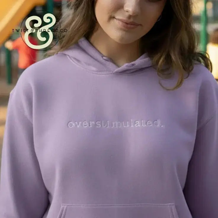 Woman wearing lavender hoodie with embroidered 'overstimulated' text in outdoor park setting
