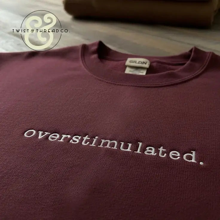 Maroon Gildan t-shirt with white embroidered 'overstimulated.' text, displayed on wooden surface