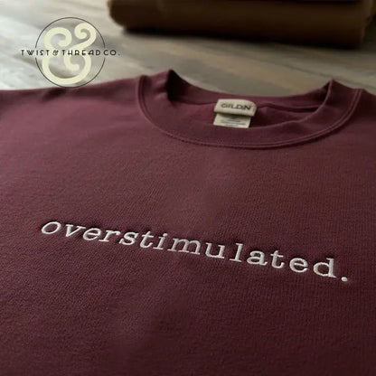 Maroon Gildan t-shirt with white embroidered 'overstimulated.' text, displayed on wooden surface