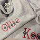 Gray dog hoodie with custom red embroidered text 'Ollie' and black paw prints, Twist & Thread Co. logo