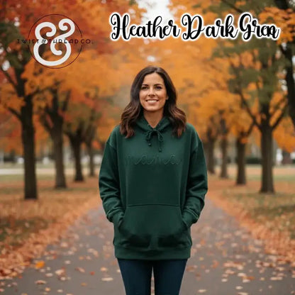 Woman wearing dark green hoodie with 'mama' embroidery standing on fall tree-lined path with orange leaves