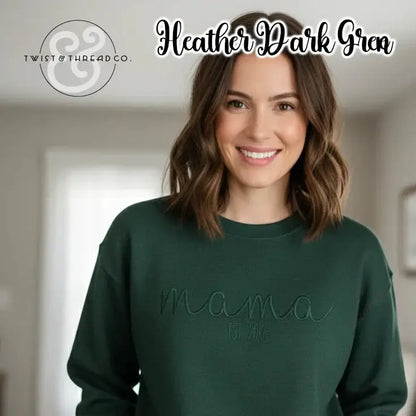Smiling woman wearing heather dark green sweatshirt embroidered with 'mama est. 2012' indoors