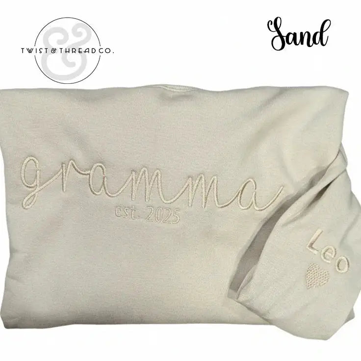Sand-colored folded sweatshirt embroidered with "gramma est. 2025" and sleeve embroidered with "Leo" and heart
