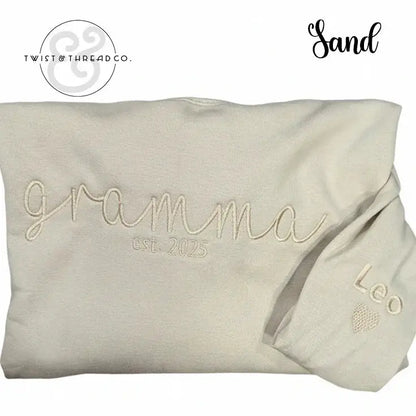 Sand-colored folded sweatshirt embroidered with "gramma est. 2025" and sleeve embroidered with "Leo" and heart