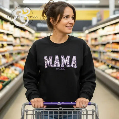 Woman wearing black sweatshirt with "MAMA est. 2012" text pushing a shopping cart in grocery store aisle