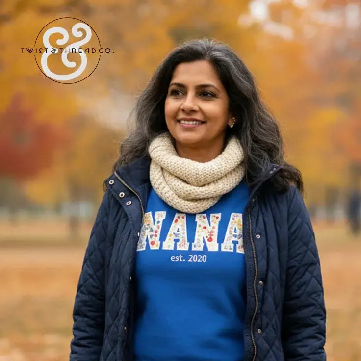 Smiling woman wearing a navy quilted jacket, cream knit scarf, and blue Nana est. 2020 sweatshirt in autumn park