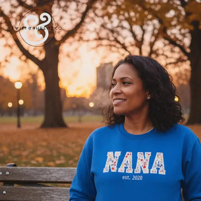 Woman wearing blue sweatshirt with floral 'NANA est. 2020' text sitting on park bench at sunset