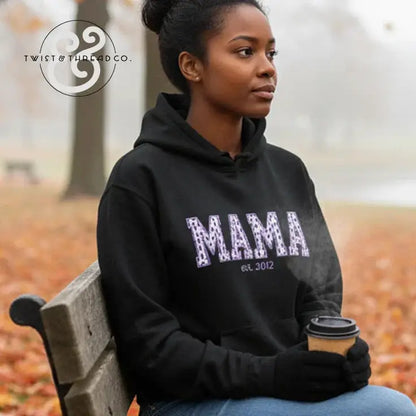 Woman wearing black hoodie with purple 'MAMA est. 3012' text, sitting on park bench holding coffee cup in fall