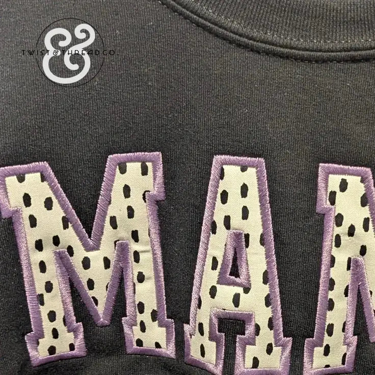 Black fabric with appliqué letters spelling 'MAM' in cream with black spots and purple stitching