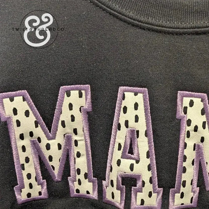 Black fabric with appliqué letters spelling 'MAM' in cream with black spots and purple stitching