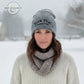 Woman wearing grey knit winter hat with embroidered text, scarf, and sweater in snowy outdoor setting