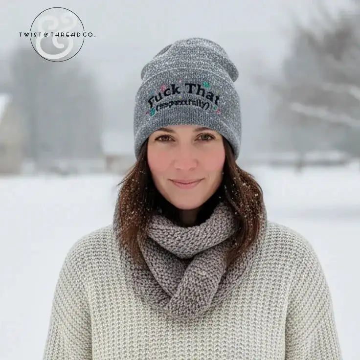 Woman wearing grey knit winter hat with embroidered text, scarf, and sweater in snowy outdoor setting