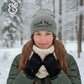 Woman in snowy forest wearing gray beanie with text, green winter jacket and cream scarf