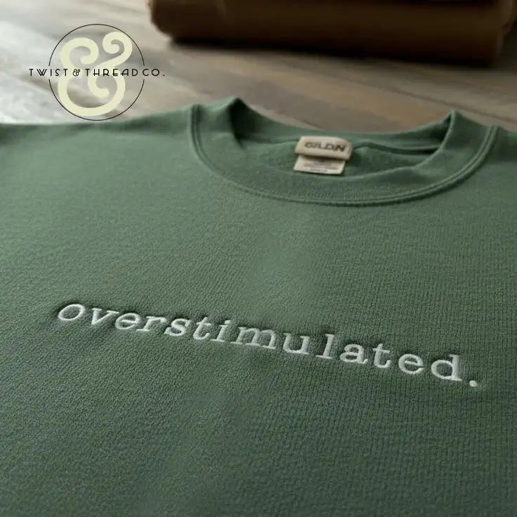 Green Gildan sweatshirt with embroidered text 'Overstimulated.' on wooden surface, Twist & Thread Co. logo