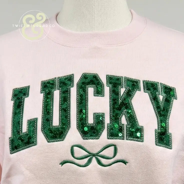 A soft pink sweatshirt featuring the word ’lucky’ in bold, green sequined lettering with a matching embroidered bow beneath it.
