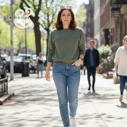 Woman walking on sunny city sidewalk wearing olive sweatshirt, blue jeans, white sneakers