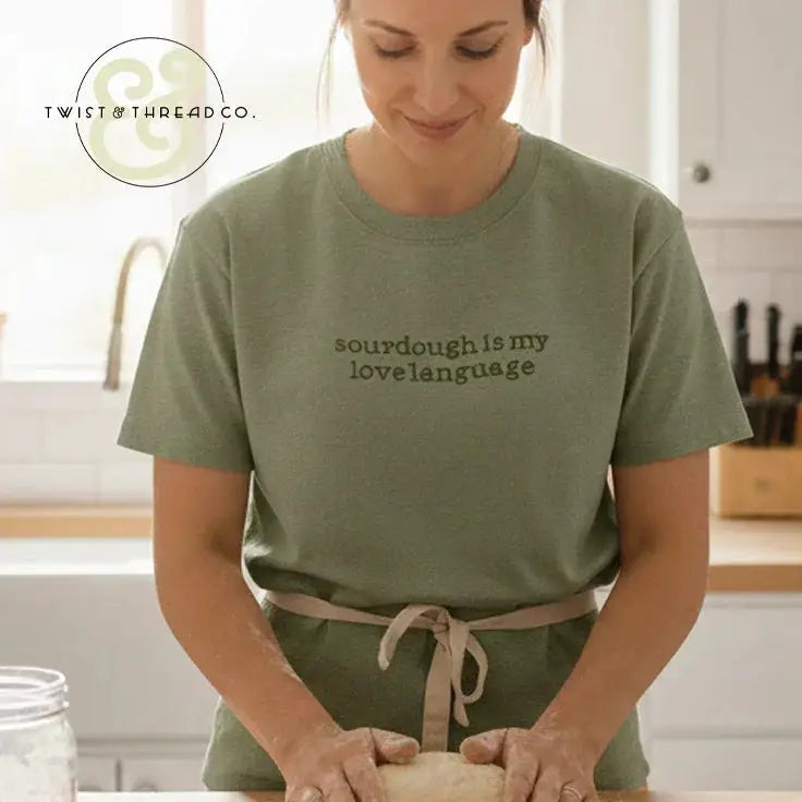 Woman in olive t-shirt with 'sourdough is my love language' text kneading dough in bright kitchen