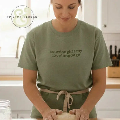 Woman in olive t-shirt with 'sourdough is my love language' text kneading dough in bright kitchen