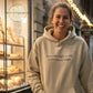 Smiling woman wearing beige hoodie with 'sourdough is my love language' text, bakery breads in background