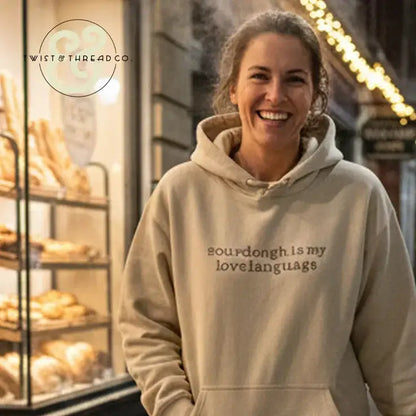 Smiling woman wearing beige hoodie with 'sourdough is my love language' text, bakery breads in background