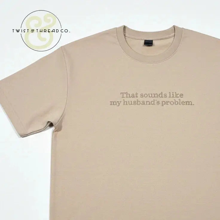Beige T-shirt with text 'That sounds like my husband's problem' printed on front, laid flat on white background