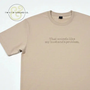 Beige T-shirt with text 'That sounds like my husband's problem' printed on front, laid flat on white background