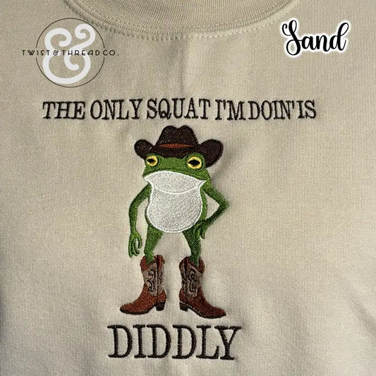Embroidered green frog in cowboy hat and boots on sand fabric with text 'The only squat I'm doin' is diddly'