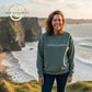 Smiling woman in custom embroidered sweatshirt reading 'Tomorrow needs you' by Twist & Thread Co. near cliffs and ocean.